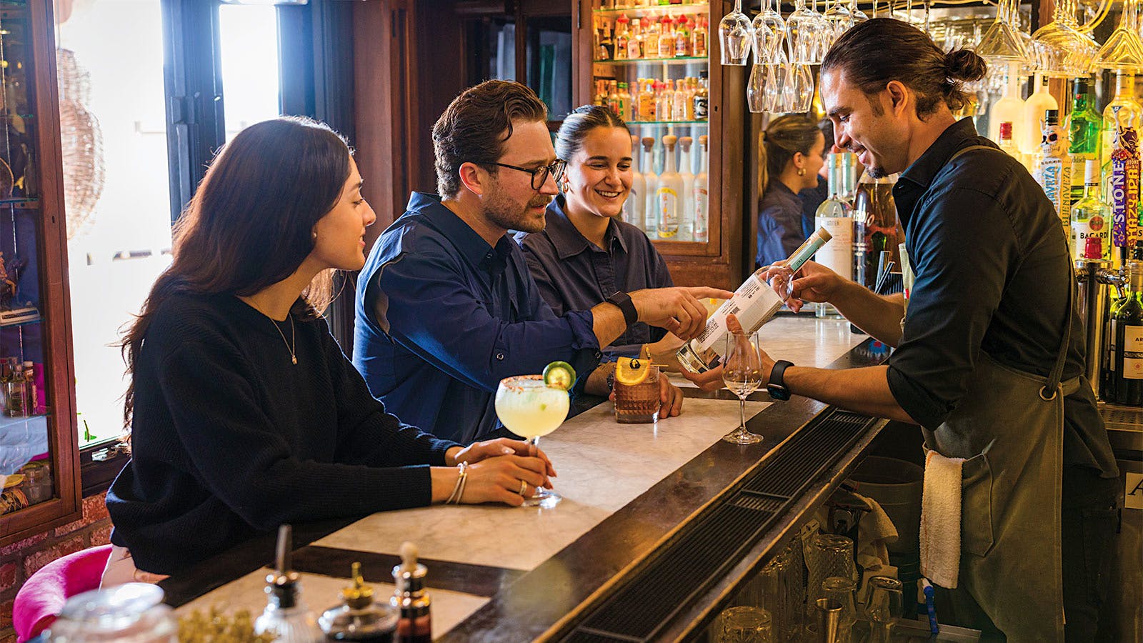 A bartender behind the bar shows a bottle of tequila to guests seated at the bar of El Agave Restaurant and Tequileria in San Diego, CA.