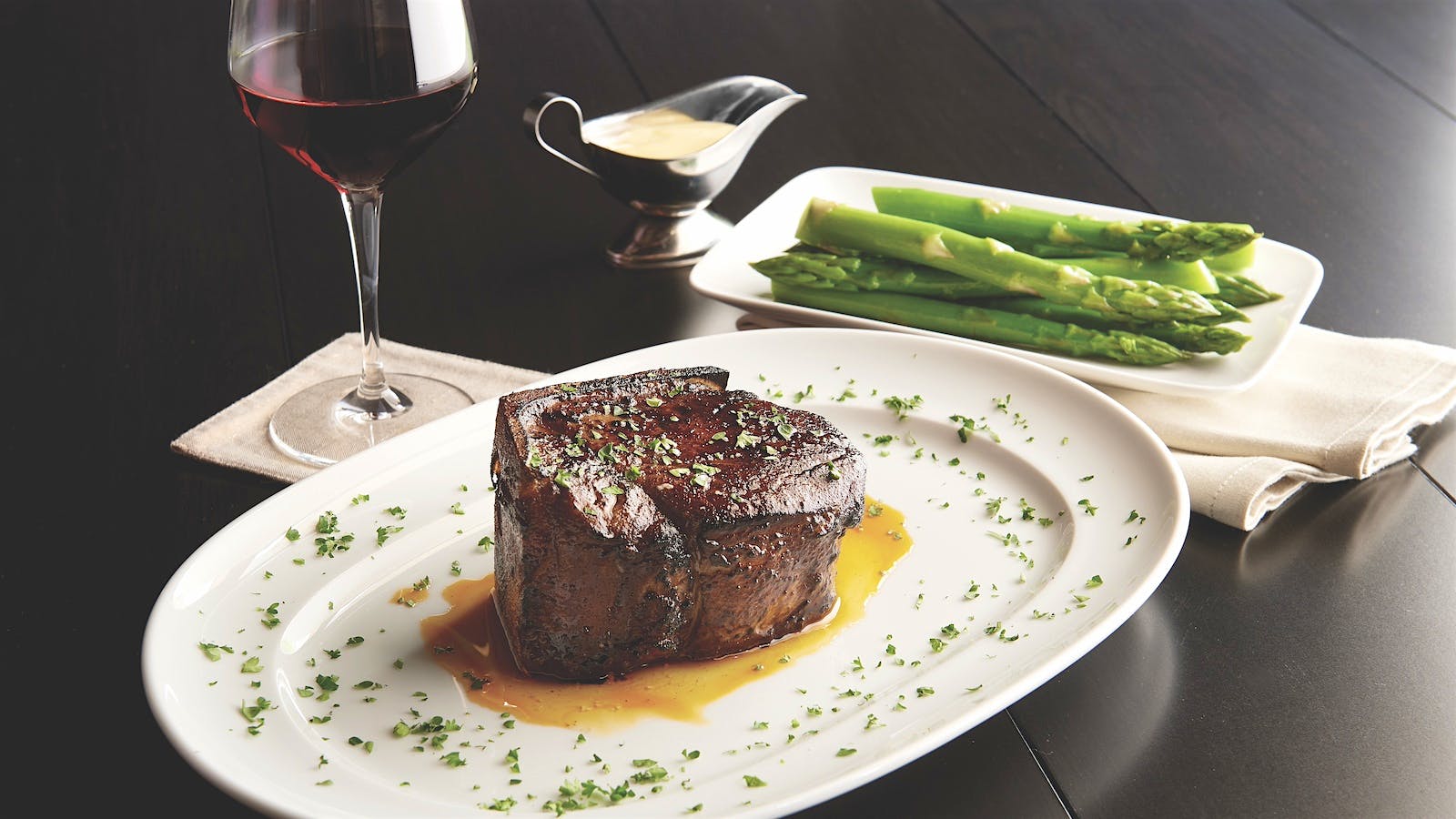 A plate of Hyōgo Kobe steak at Mastro's Steakhouse in Houston, Texas, served with a side of asparagus, pictured next to a gravy boat with white sauce and a glass of red wine.