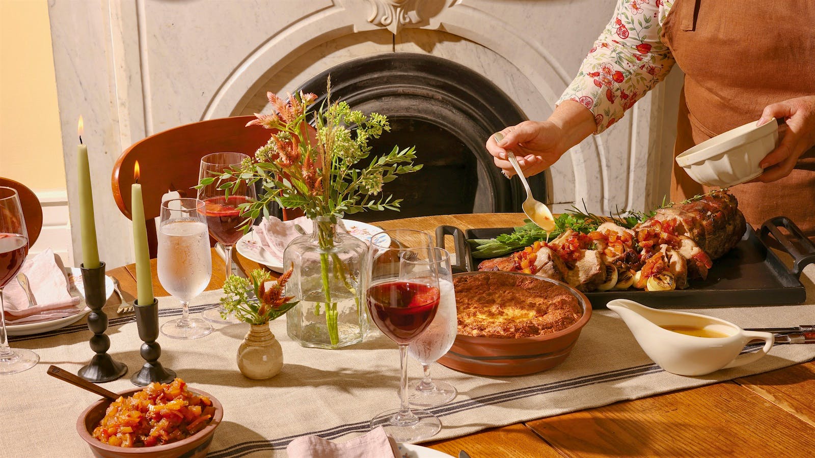 Ouita Michel drizzles gravy over a cast iron serving dish of sliced Bourbon-brined pork roast, garnished with tipsy fruit chutney, rosemary and sage, alongside a dish of cheese grits soufflé and a gravy boat.