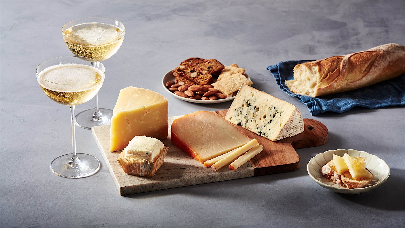 A plate of four cheeses alongside two glasses of sparkling wine and a baguette